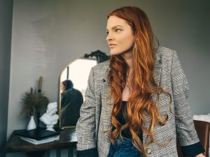 Redhead woman with long hair leaning against a desk wearing a sport coat and jeans.